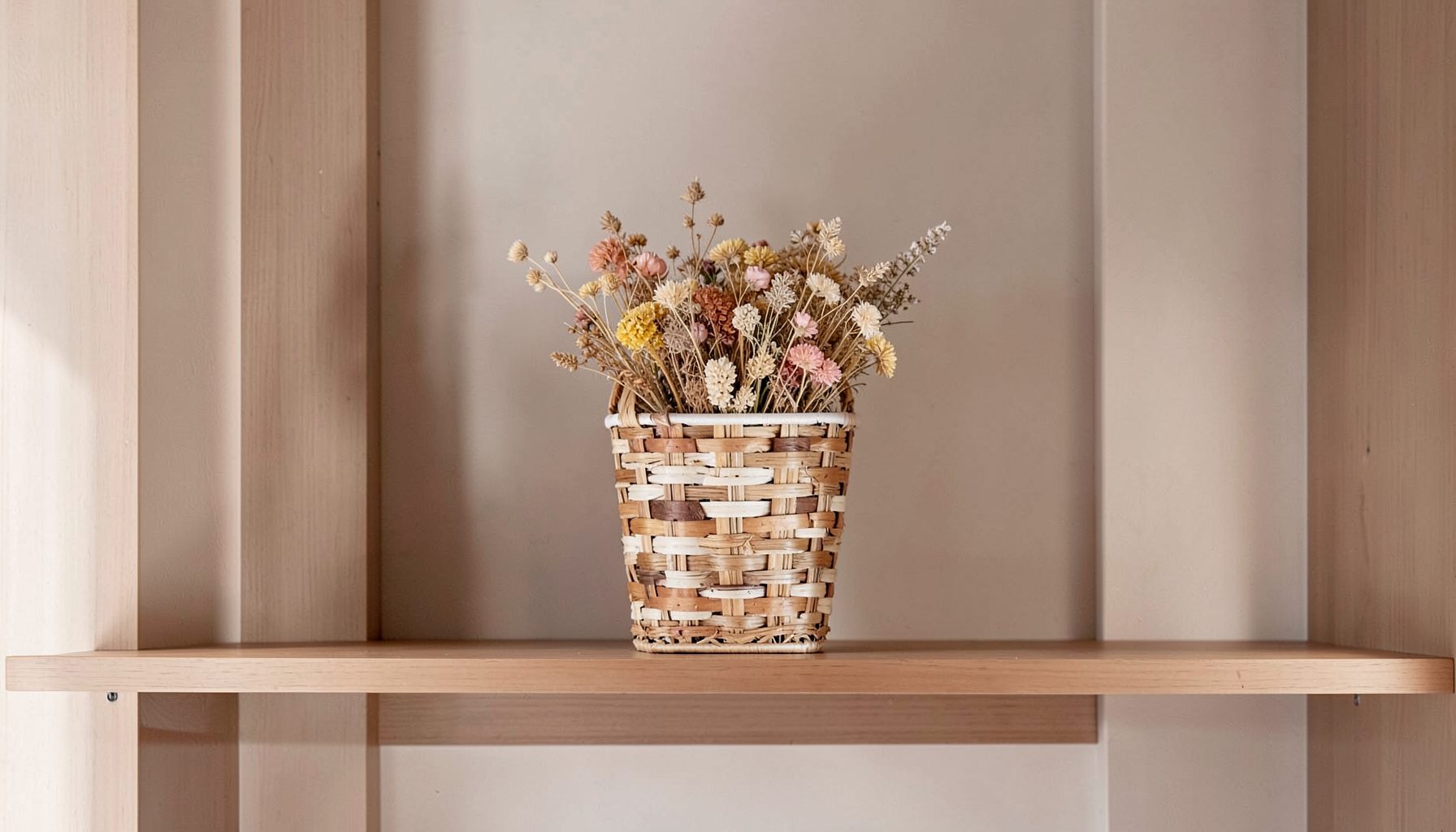 A decorative basket woven from strips of a repurposed ice cream pail, holding dried flowers on a wooden shelf