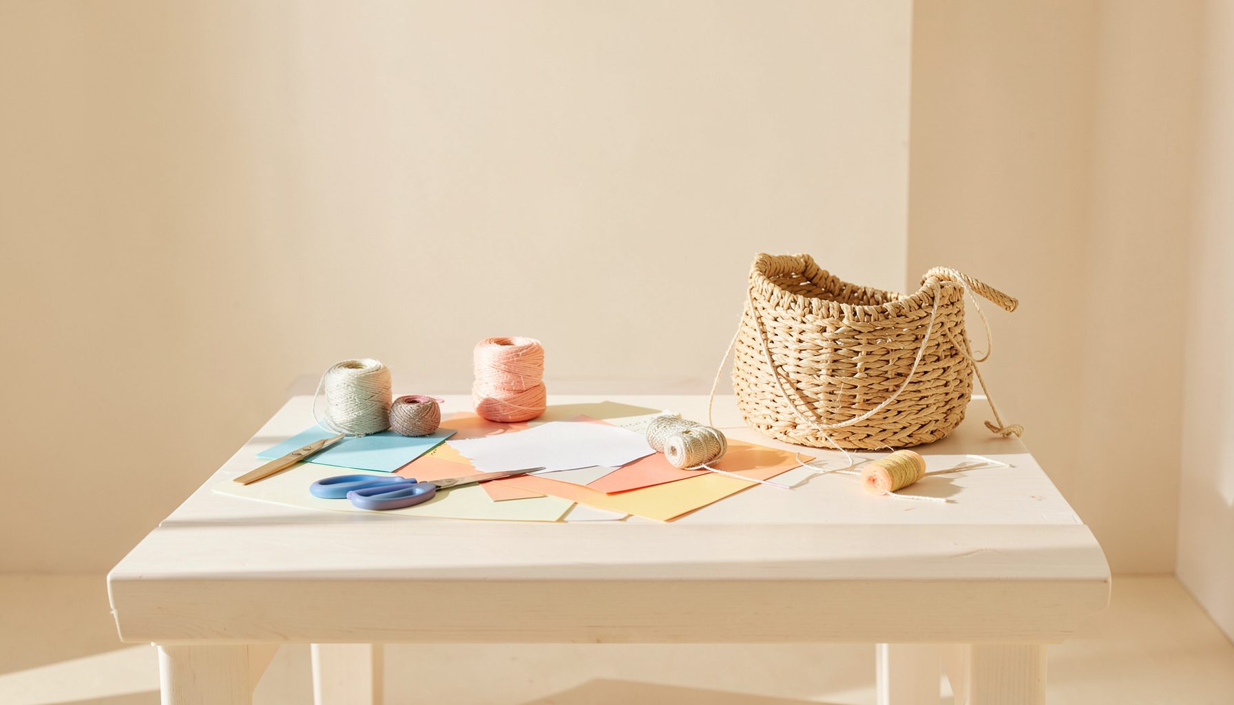 A bright, airy craft table with scissors, colored paper, yarn spools, and a half-finished woven basket in warm natural light, soft beige and cream tones