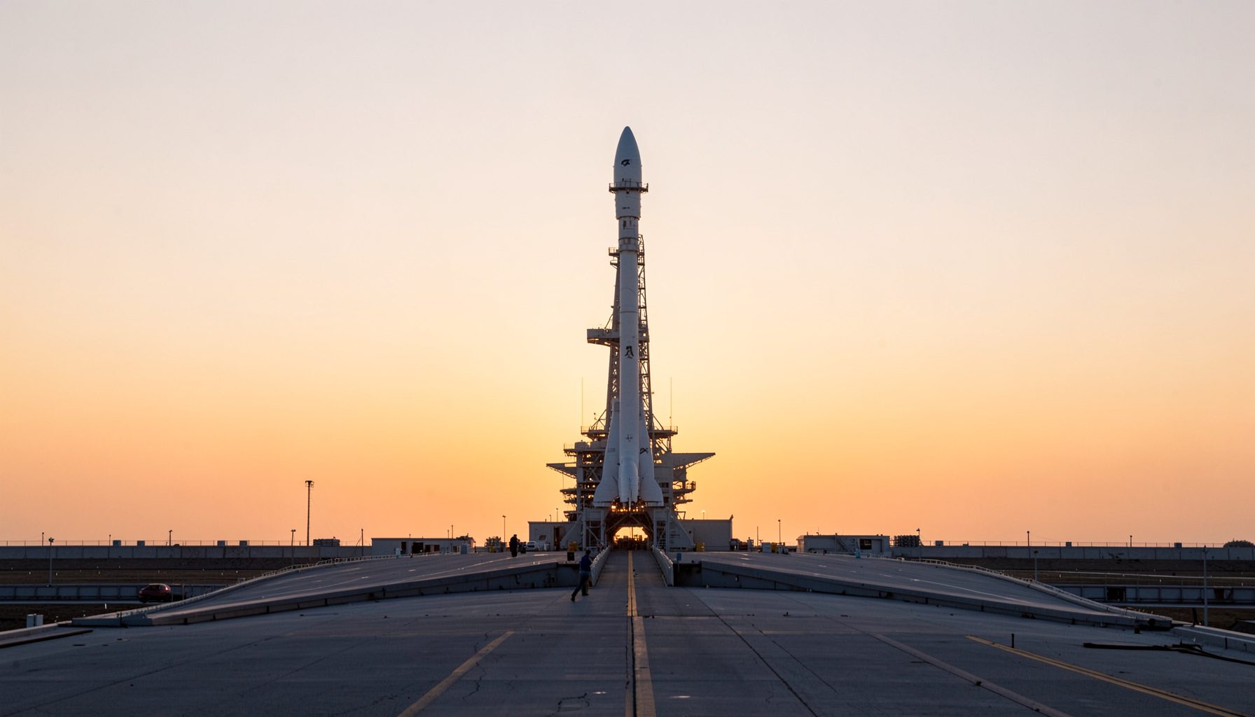 A clean architectural photograph of a NASA launch pad at dawn, with strong symmetry and a rocket silhouetted against a pale orange sky