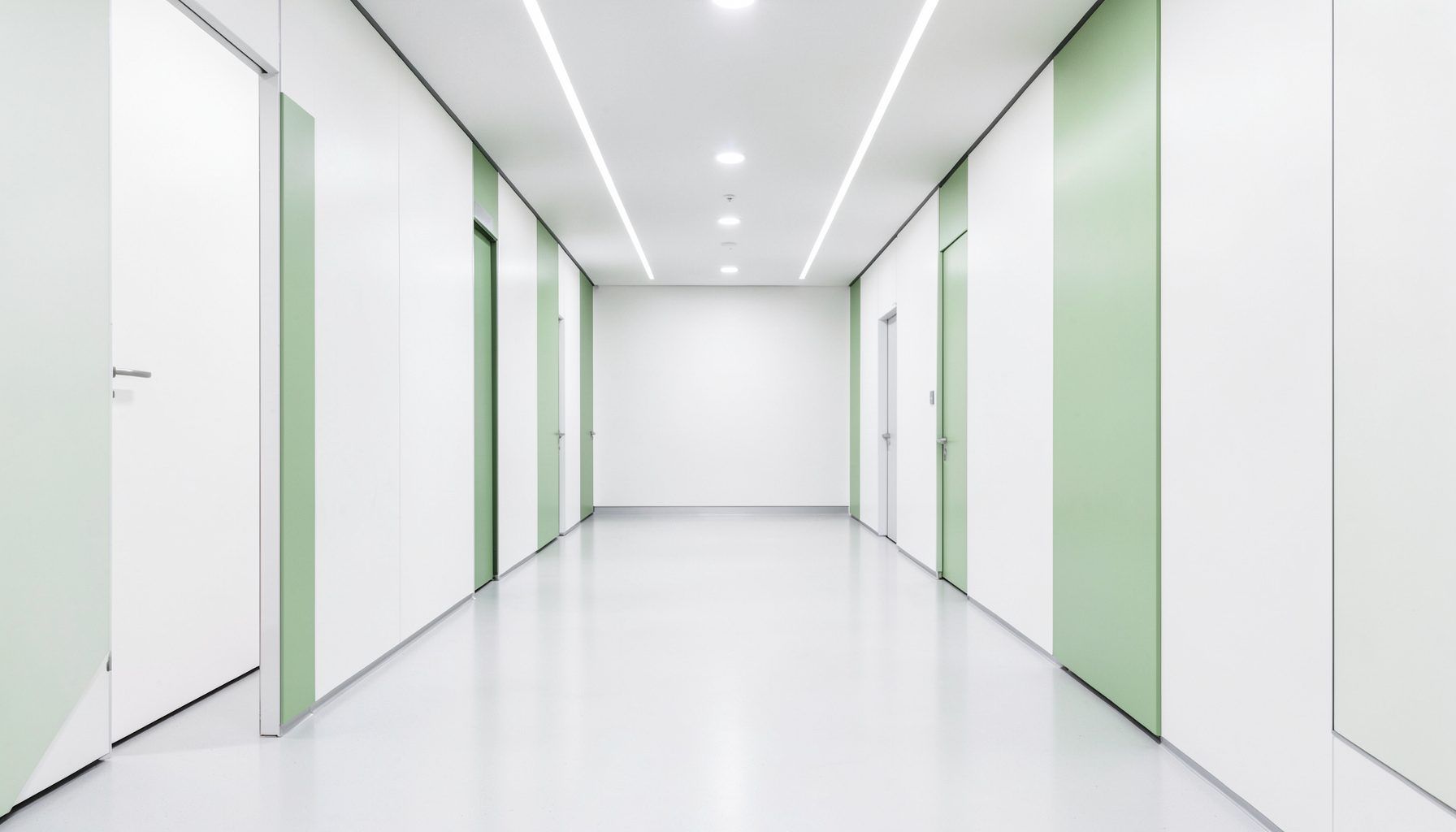 A clean architectural photograph of a futuristic medical facility corridor with strong symmetry, white walls, and soft green accent panels