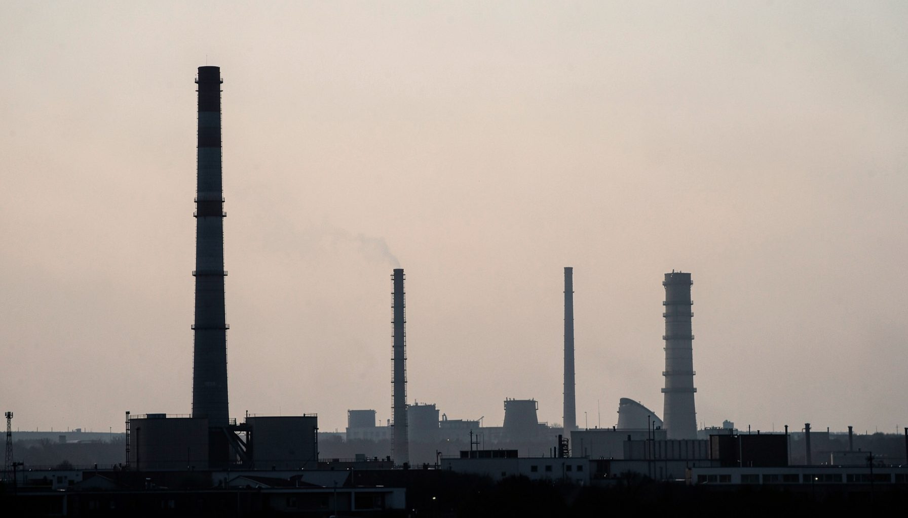 A stark industrial landscape with smokestacks and cooling towers silhouetted against a hazy sky, rendered in muted grays and pale orange.