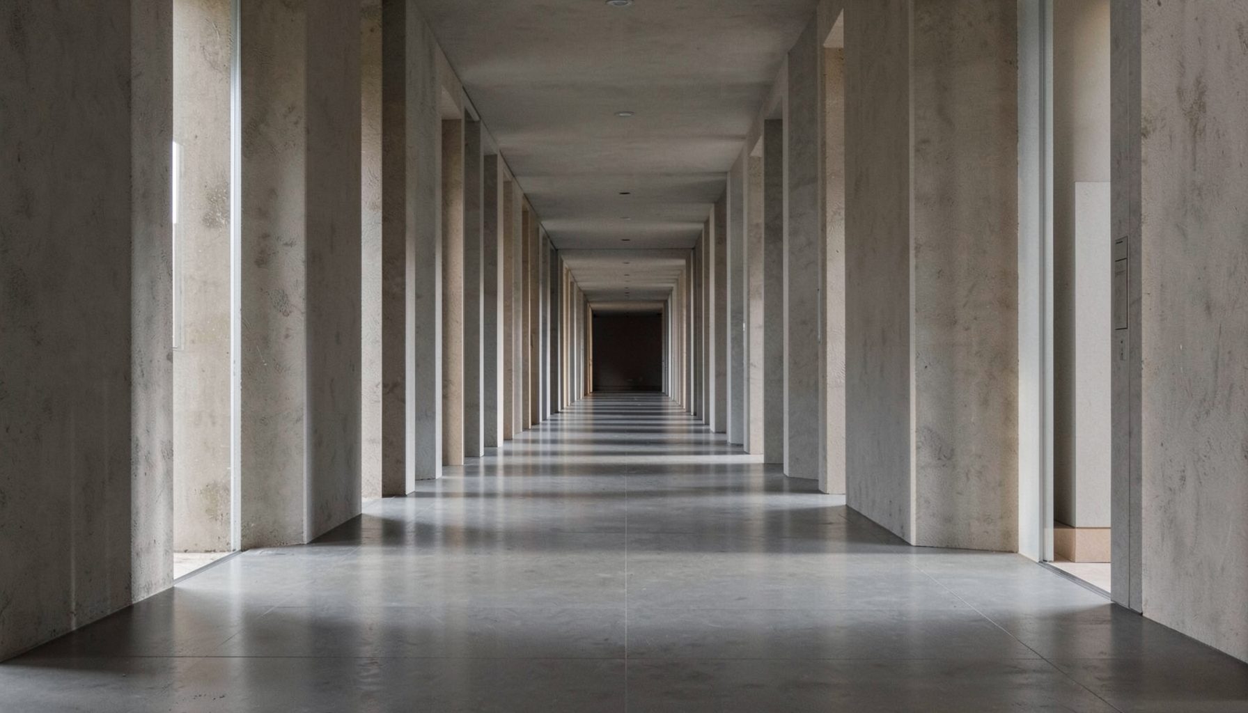 A clean architectural photograph with strong symmetry: a long corridor or colonnade receding into the distance, bathed in soft natural light with deep shadows, rendered in muted grays and warm neutrals.
