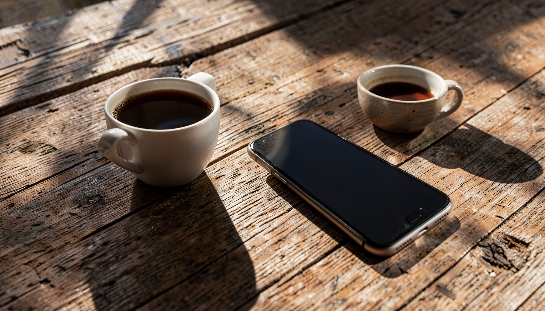 A smartphone resting on a weathered wooden table beside a half-empty coffee cup, morning light casting long shadows, in warm browns and soft whites.
