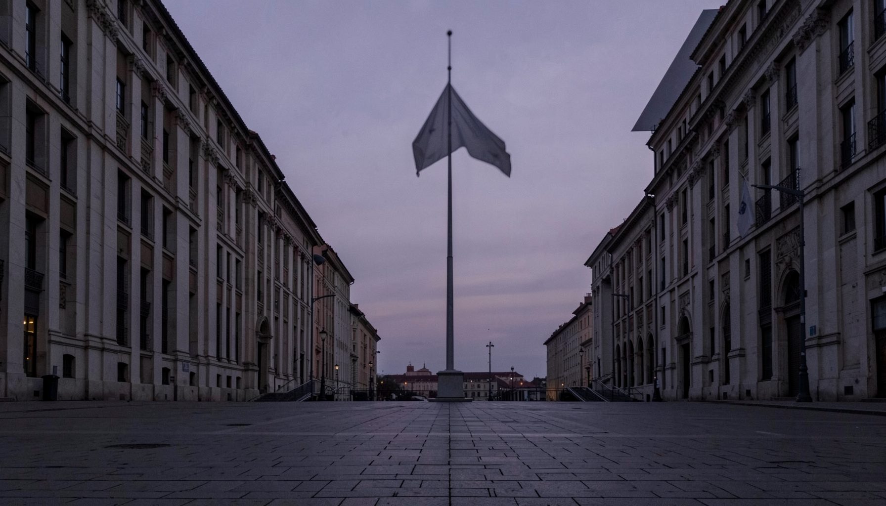 A quiet public square at dusk with a flag at half-mast, soft purple and gray sky, conveying solemnity and reflection.