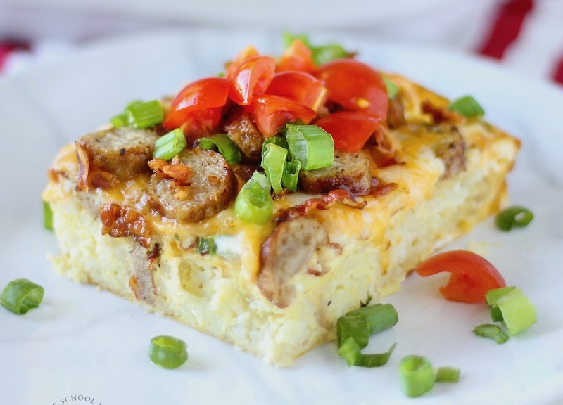Slice of a baked egg casserole topped with sausage, cheese, diced tomatoes, and green onions on a white plate